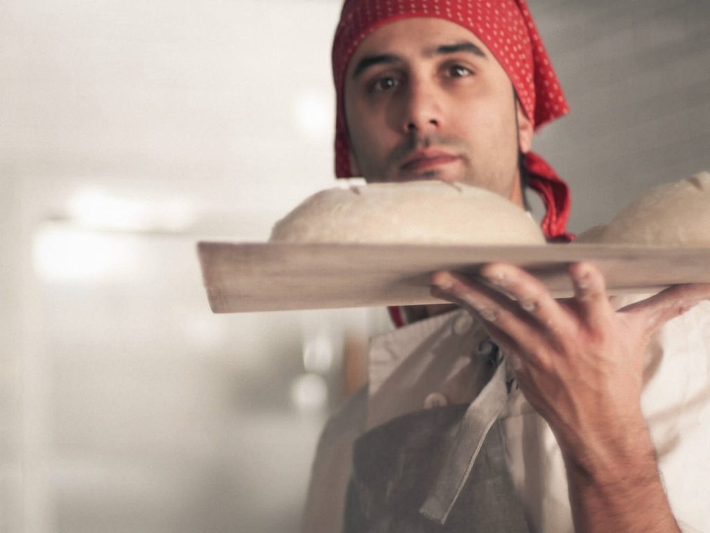 Memoria audiovisual de un pequeño negocio: bandeja con hogazas de pan en primer plano y el panadero desenfocado al fondo.